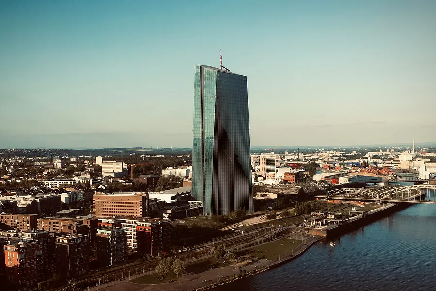 Vista aérea del imponente edificio del Banco Central Europeo (BCE) en Frankfurt, Alemania. La estructura de vidrio y acero se eleva majestuosa sobre el horizonte urbano, rodeada de edificios modernos y tradicionales. En primer plano, el río Main serpentea junto a una zona verde, añadiendo un toque natural a la escena. El cielo está despejado, resaltando la silueta del rascacielos contra el fondo azul.