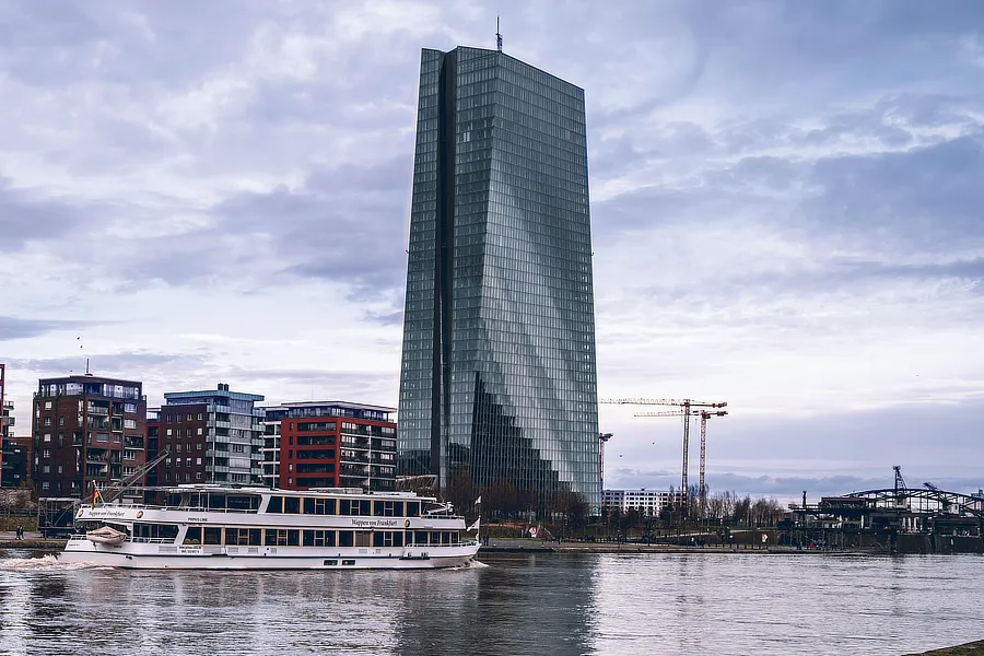 El edificio del Banco Central Europeo (BCE) se alza imponente junto al río Meno en Frankfurt, con un cielo nublado de fondo. Un barco blanco navega por el río, mientras grúas y edificios modernos rodean la escena.