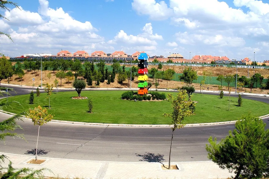 La imagen muestra una rotonda en Boadilla del Monte, adornada con una colorida escultura de cubos apilados. Alrededor, se observa un área verde bien cuidada con árboles pequeños y arbustos, y al fondo, un barrio residencial con casas de techos rojizos. El cielo azul y las nubes blancas añaden un toque de serenidad al paisaje.