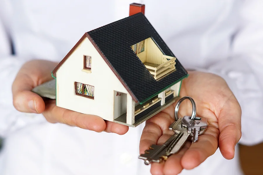 Se ve medio tronco de una persona con camisa blanca. Y en primer plano sus dos manos. Con la mano derecha sostiene la maqueta de una casa con fachada blanca y techo azul oscuro y en la palma de su mano izquierda muestra las llaves de una vivienda.