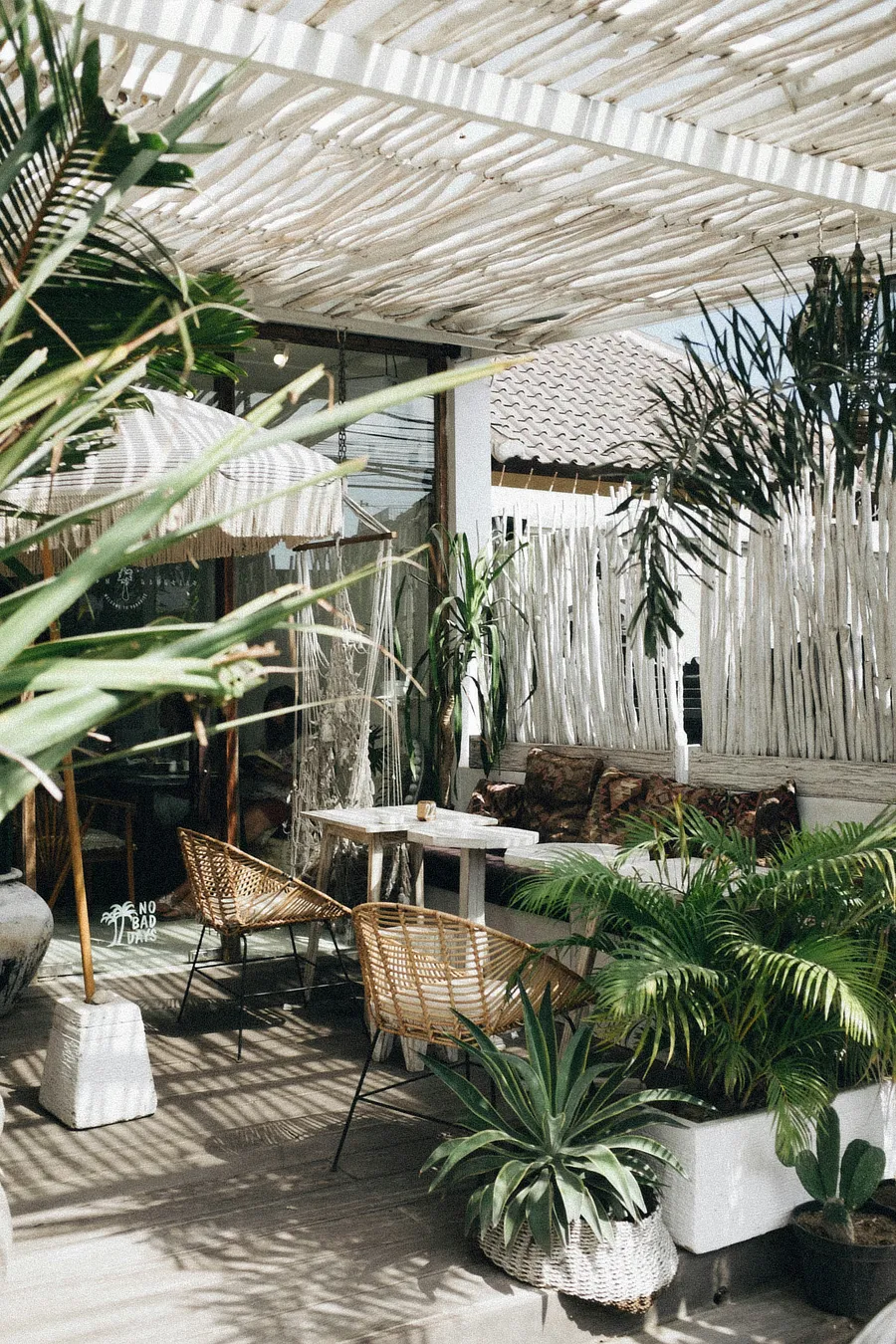 Imagen donde se ve una terraza entre plantas. La terraza tiene un techado de madera en color claro, una sombrilla con pie blanco, mesa y sillas.