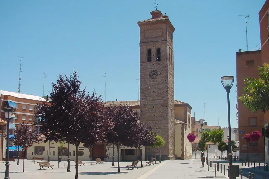 Vista de la torre de la Iglesia de Nuestra Señora de la Asunción en Móstoles, con árboles y bancos en la plaza circundante, destacando un entorno urbano tranquilo y bien cuidado