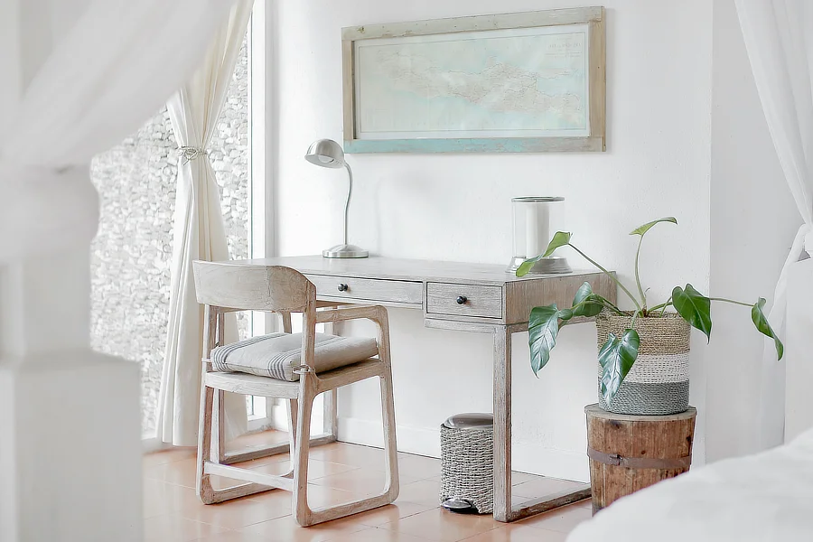 Un elegante espacio de teletrabajo con una decoración minimalista. Se observa una mesa de madera clara con dos cajones, una silla acolchada y un flexo plateado. La mesa está situada junto a una gran ventana que proporciona abundante luz natural. A la derecha de la mesa, hay una planta en una maceta decorativa y un cuadro en la pared con un mapa antiguo.