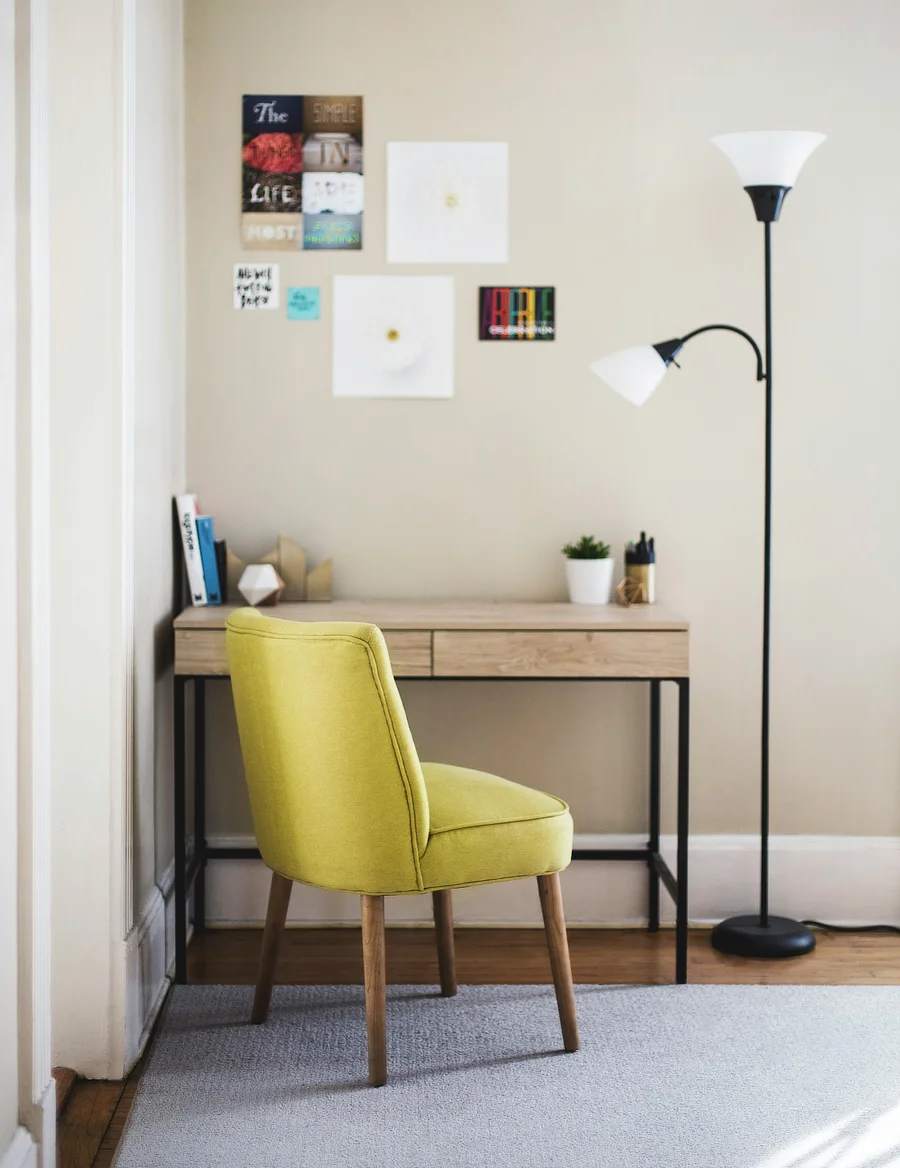 Un acogedor rincón de teletrabajo con una decoración moderna y colorida. Una mesa de madera clara con patas negras sostiene algunos objetos decorativos y papelería. Frente a la mesa, hay una silla de color verde oliva con tapizado acolchado. En la pared sobre la mesa, se ven varios cuadros pequeños y coloridos. A la derecha, una lámpara de pie con dos luces proporciona iluminación adicional al espacio.