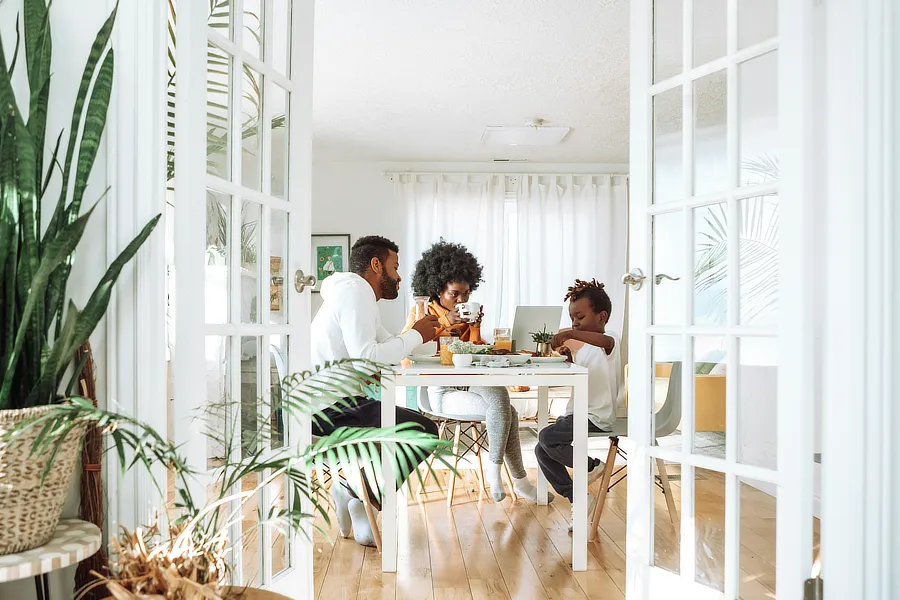 Una familia desayuna con su hijo de unos 6-8 años en el salón de su casa. Un salón en tonos claros, con puertas blancas. Mesa y silla en color blanco. El suelo es de parqué claro.