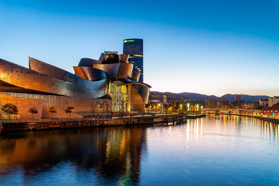 Vista de la caída del sol sobre la ría de Bilbao con el Museo Guggenheim a la izquierda de la foto, la parte derecha la ocupa completamente la ría. Con la ciudad al fondo de la foto bajo un cielo limpio azul que se oscurece por la llegada de la noche.