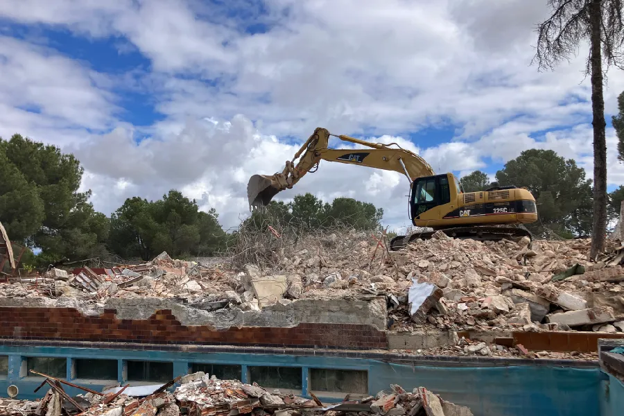 La imagen muestra el proceso de demolición de la vivienda que hay en la parcela. En el centro de la fotografía, se observa una excavadora amarilla de la marca CAT, modelo 325C, trabajando sobre un gran montón de escombros. La excavadora está utilizando su brazo hidráulico para mover los restos de la edificación derrumbada. En primer plano se puede ver una piscina parcialmente demolida con restos de ladrillos, cemento y otros materiales de construcción esparcidos alrededor. La piscina, de color azul, contrasta con los tonos grises y marrones de los escombros y los ladrillos. Al fondo de la foto, se aprecian árboles verdes que rodean el sitio de demolición, bajo un cielo parcialmente nublado con secciones de cielo azul visible. La imagen captura el momento activo de la demolición, mostrando tanto la maquinaria en acción como los restos de la estructura demolida, destacando el avance en la preparación del terreno para la futura construcción de los 8 chalets de lujo.