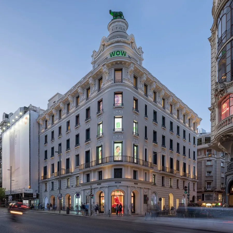 La imagen muestra una vista del edificio WOW en la Gran Vía de Madrid. Este icónico edificio de estilo neoclásico se destaca por su elegante arquitectura y su ubicación estratégica en una de las principales avenidas comerciales de la ciudad. En la imagen, se puede ver la fachada del edificio iluminada al atardecer, con escaparates llamativos que exhiben productos y pantallas digitales que atraen a los transeúntes. El tráfico y los peatones añaden dinamismo a la escena urbana, mientras que el rótulo "WOW" en la parte superior del edificio indica la presencia de las modernas galerías comerciales que alberga en su interior.