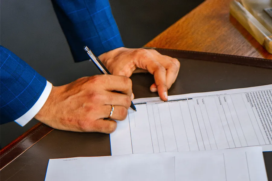 Persona firmando un documento con un bolígrafo en una mesa, representando la firma de una nota de encargo en el proceso inmobiliario. La persona viste una chaqueta azul y la mesa es de madera oscura, con documentos y un bolígrafo en la superficie.