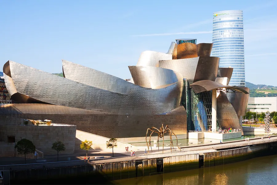 Vista del Museo Guggenheim Bilbao durante el día, destacando su arquitectura moderna con paneles de titanio curvados y brillantes. En primer plano, la famosa escultura de araña de Louise Bourgeois, 'Maman', se encuentra junto al río. Al fondo, se puede ver la torre Iberdrola, un rascacielos que forma parte del paisaje urbano de Bilbao.