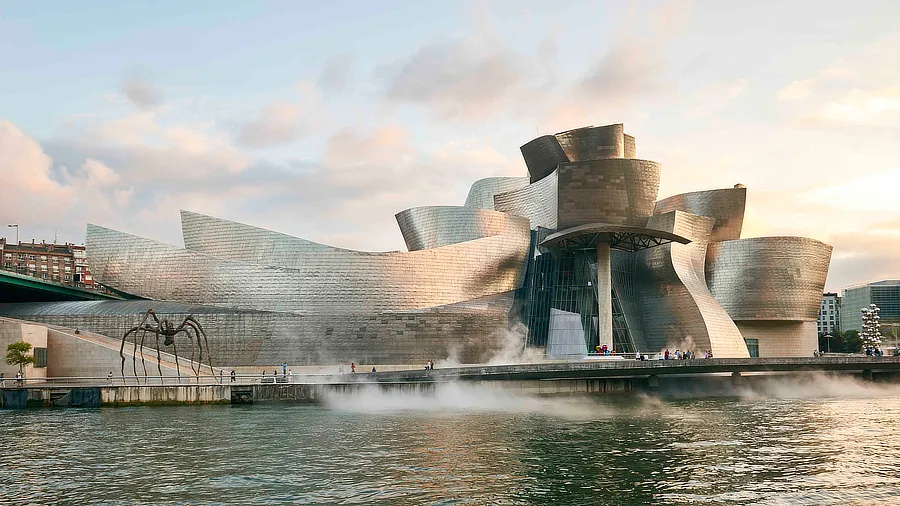 Vista del Museo Guggenheim Bilbao al atardecer, destacando su arquitectura moderna con paneles de titanio curvados y brillantes. En primer plano, el río refleja la estructura, mientras que la famosa escultura de araña de Louise Bourgeois, 'Maman', se observa a la izquierda.