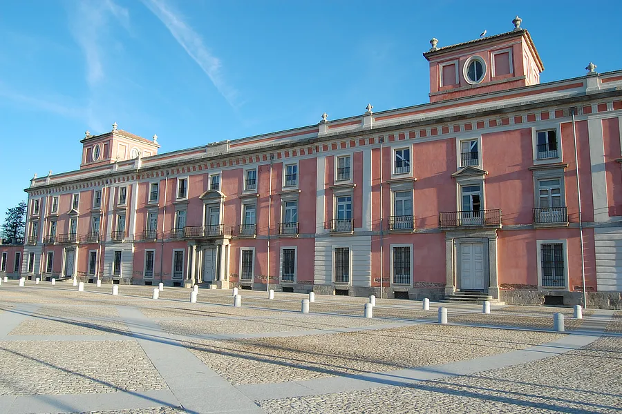 Fachada Principal del Palacio. La imagen muestra la fachada principal del Palacio del Infante don Luis en Boadilla del Monte. Se observa su diseño neoclásico con simetría perfecta, incluyendo balcones franceses, pilastras y una entrada flanqueada por columnas dóricas. La fachada de color rosado contrasta con el cielo azul, destacando la elegancia y majestuosidad del edificio.