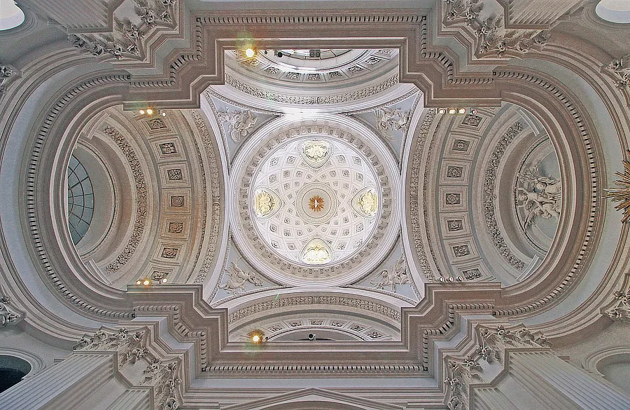 Cúpula de la Capilla. La imagen muestra la impresionante cúpula de la capilla del Palacio del Infante don Luis. Se observa el intrincado diseño arquitectónico con detalles ornamentales en relieve y motivos florales. La cúpula está adornada con esculturas de ángeles y otros elementos decorativos, iluminados por la luz natural que entra a través de las ventanas altas, creando un efecto de luminosidad y grandeza.