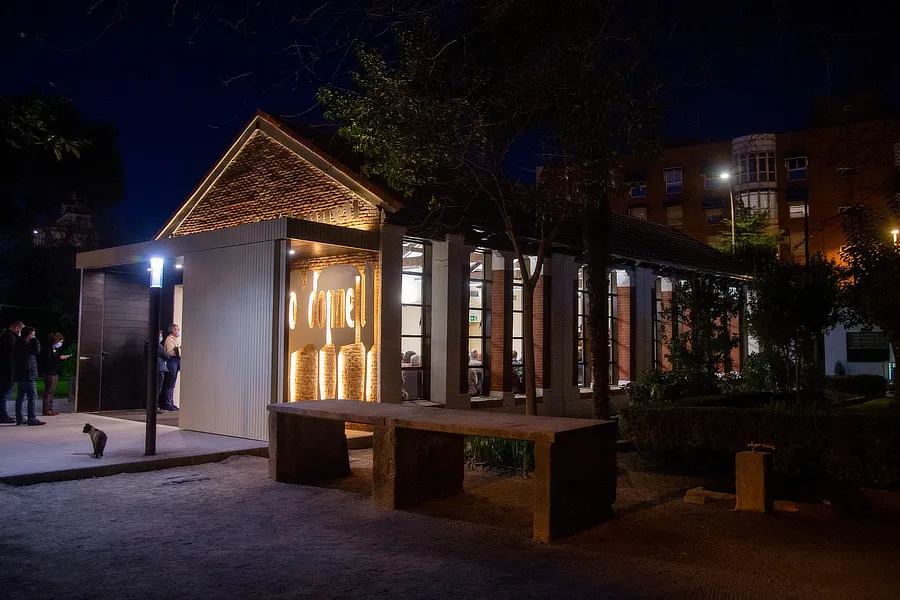 Vista nocturna de la Casita del Parque O'Donnell en Alcalá de Henares, iluminada con luces cálidas que resaltan su arquitectura. Alrededor, se ven algunos visitantes y un gato en primer plano, con un fondo de edificios y árboles.