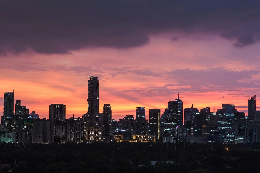 Vista panorámica del horizonte de Manila al atardecer, con edificios altos y modernos reflejándose en el agua. El cielo está pintado en tonos rosados y azulados, creando una atmósfera serena y elegante. La imagen captura la esencia de una ciudad vibrante y moderna, destacando la belleza arquitectónica y la tranquilidad del momento.