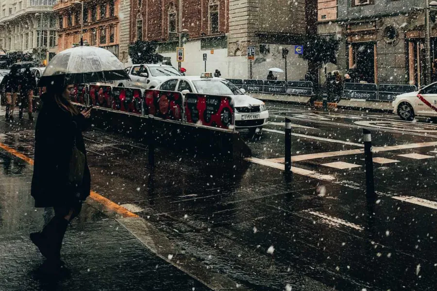 Imagen de una calle de Madrid donde se ven algunos taxis y una persona en la parte izquierda llevando un paraguas abierto para protegerse de la nieve que está cayendo.