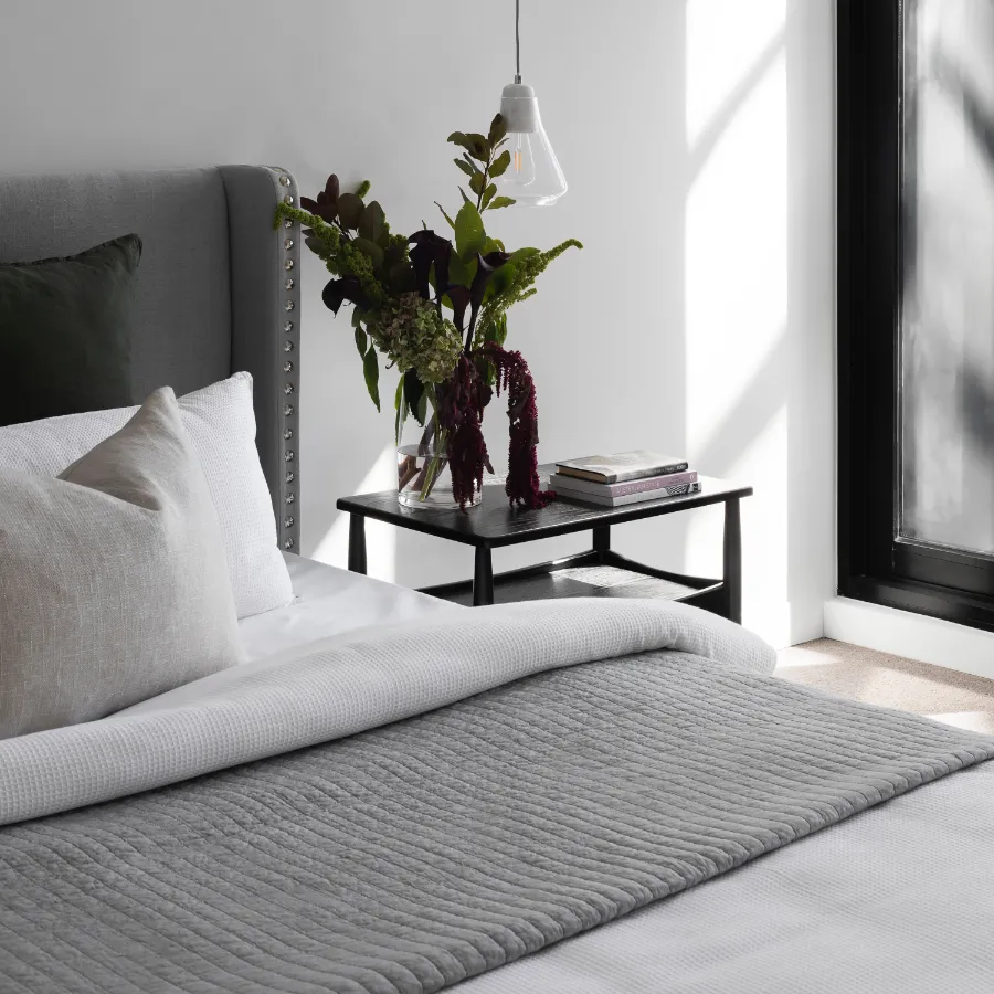 Vista parcial de una cama con colcha de color ocre y manta más oscura con una bonita mesa de noche.