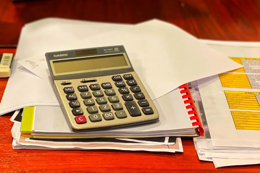 Sobre una mesa de color cedro hay dos pilas de documentos dina A4 encanutillados y sobre el montón de la izquierda una calculadora solar Casio.