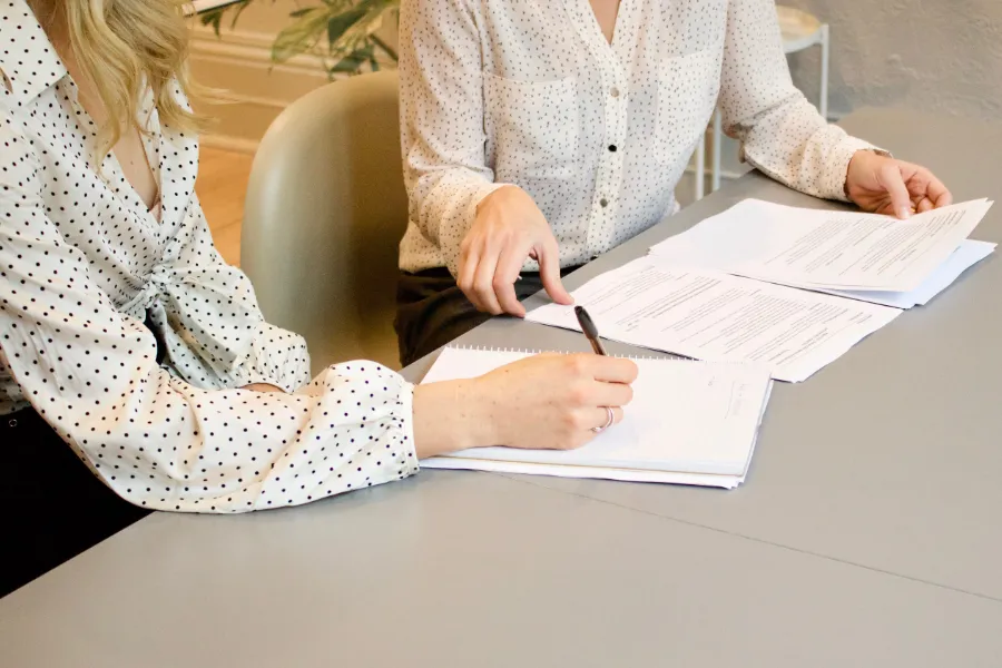Dos señoras de las que solo se ve medio torso están sentadas frente a una mesa con una superficie grisácea revisando unas notas sobre lo que parece un contrato.