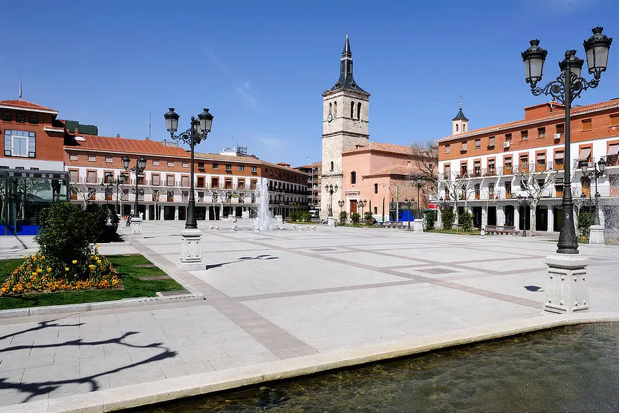 En la imagen se puede ver la Plaza Mayor de Torrejón de Ardoz. En el centro destaca una fuente rodeada de bancos y flores. Al fondo, la iglesia de San Juan Evangelista con su campanario. La plaza está flanqueada por edificios de estilo tradicional, lámparas de hierro y una zona peatonal amplia y limpia.