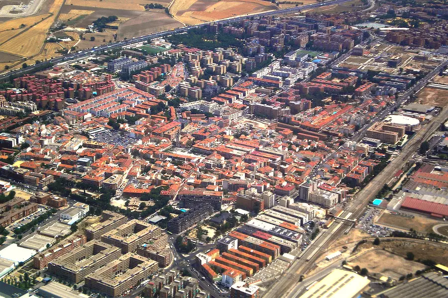 En la imagen se muestra una vista aérea de Torrejón de Ardoz. Se pueden apreciar múltiples edificios residenciales con tejados rojos, organizados en bloques. También se observan carreteras y vías ferroviarias que atraviesan la ciudad, así como áreas industriales y campos en los alrededores, reflejando la mezcla urbana y rural del municipio.